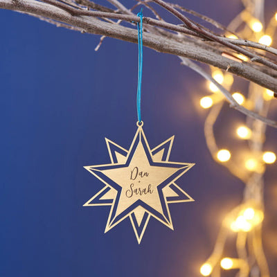 Star-shaped ornament with 'Dan + Sarah' engraving hanging on a branch with blurred lights in the background